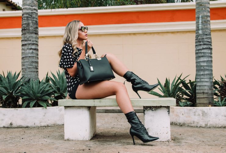 woman wearing black and white polka dot shirt with black short shorts holding black leather tote bag sitting on white concrete bench