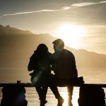 silhouette of man and woman sitting on bench near body of water during sunset