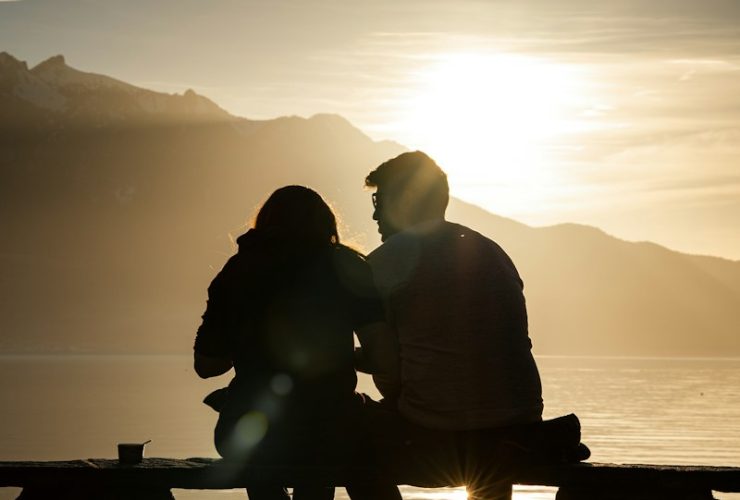 silhouette of man and woman sitting on bench near body of water during sunset