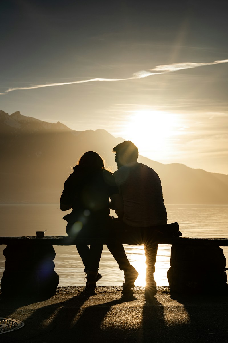 silhouette of man and woman sitting on bench near body of water during sunset