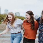 three women walking on brown wooden dock near high rise building during daytime