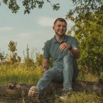 man in green polo shirt and blue denim jeans sitting on brown wooden log during daytime