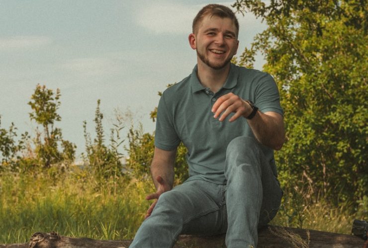 man in green polo shirt and blue denim jeans sitting on brown wooden log during daytime