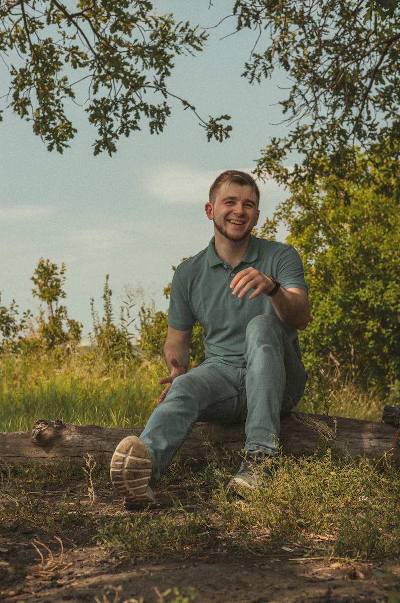 man in green polo shirt and blue denim jeans sitting on brown wooden log during daytime